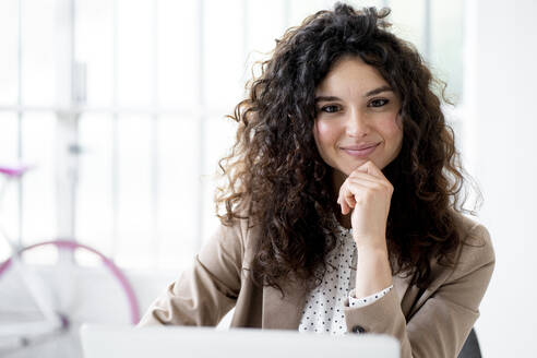 Lächelnde Geschäftsfrau mit Hand am Kinn, die im Büro sitzt - GIOF13097
