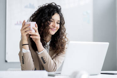 Businesswoman with coffee cup smiling while looking at laptop - GIOF13106