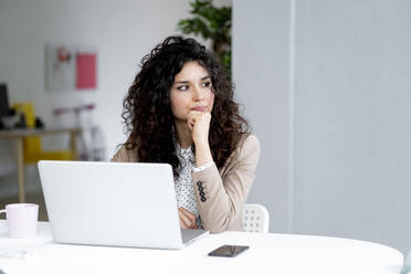 Businesswoman looking away while sitting with hand on chin at desk in office - GIOF13108