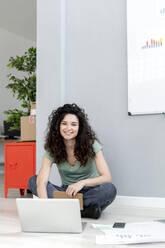 Lächelnde Geschäftsfrau sitzt im Schneidersitz auf dem Boden im Büro - GIOF13115