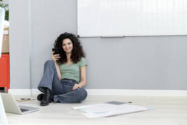 Lächelnde Geschäftsfrau mit Smartphone an die Wand gelehnt, während sie im Büro auf dem Boden sitzt - GIOF13119