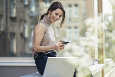Lächelnde Geschäftsfrau, die ein Mobiltelefon benutzt, während sie mit einem Laptop auf dem Sitz sitzt - UUF24331