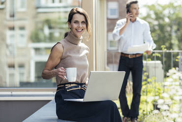 Smiling businesswoman holding coffee cup while sitting with colleague in background - UUF24335