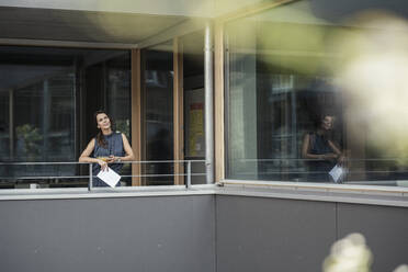 Geschäftsfrau mit digitalem Tablet, die auf dem Balkon eines Büros steht und nachdenkt - UUF24364