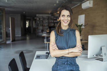 Smiling businesswoman with arm crossed leaning on desk - UUF24381