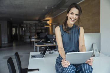 Fröhliche Geschäftsfrau mit digitalem Tablet auf dem Schreibtisch sitzend - UUF24384