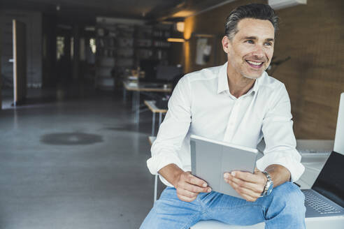 Lächelnder Geschäftsmann mit digitalem Tablet im Büro sitzend - UUF24387