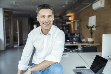 Smiling businessman sitting by laptop in office - UUF24390