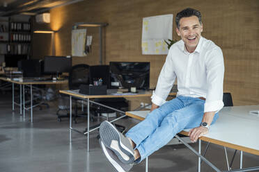 Cheerful businessman with legs crossed at ankle sitting on desk - UUF24392