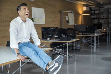 Happy businessman sitting with legs crossed at ankle on desk - UUF24393