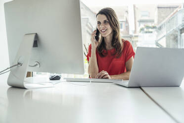 Geschäftsfrau, die mit einem Mobiltelefon spricht, während sie einen Computer im Büro benutzt - UUF24402