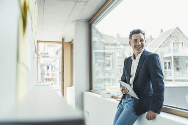 Lächelnder reifer Geschäftsmann, der ein digitales Tablet hält, während er sich auf die Fensterbank lehnt - UUF24419