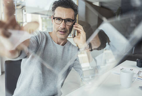 Männlicher Fachmann, der durch einen Glaskasten schaut, während er mit einem Mobiltelefon im Büro spricht - UUF24435