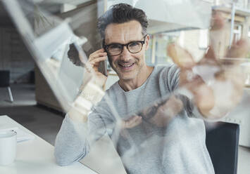 Lächelnder Geschäftsmann mit Glaskasten, der im Büro mit einem Mobiltelefon spricht - UUF24436