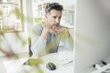 Geschäftsmann mit Händen am Kinn bei der Arbeit am Computer im Büro - UUF24440