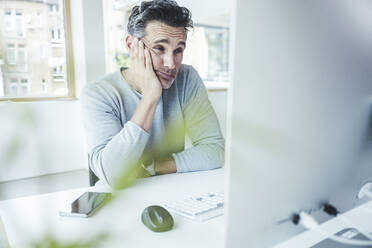 Müder Geschäftsmann mit Hand am Kinn bei der Arbeit am Computer im Büro - UUF24441