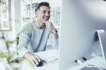 Lächelnder reifer Geschäftsmann, der einen Computer benutzt, während er im Büro sitzt - UUF24442