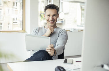 Lächelnder Geschäftsmann mit digitalem Tablet im Büro sitzend - UUF24444
