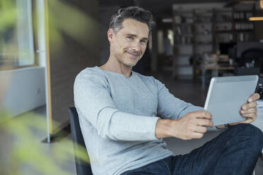 Lächelnder reifer Geschäftsmann, der ein digitales Tablet hält, während er am Schreibtisch sitzt - UUF24448