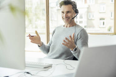 Businessman with headset gesturing while attending video call through computer in office - UUF24449