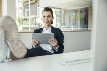 Weibliche Fachkraft, die ein digitales Tablet hält und ein Bein auf dem Schreibtisch abstützt - UUF24461