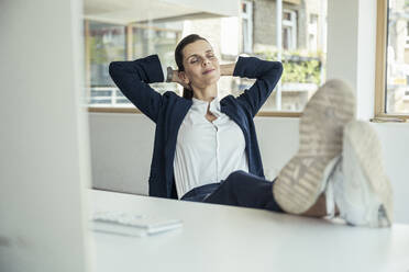 Lächelnde Geschäftsfrau mit Händen hinter dem Kopf, die sich im Büro entspannt - UUF24465