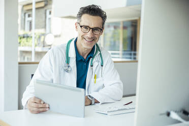 Lächelnder männlicher Arzt mit digitalem Tablet im Büro - UUF24468