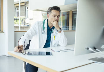 Male doctor with eyes closed and hand on chin sitting at desk in office - UUF24471