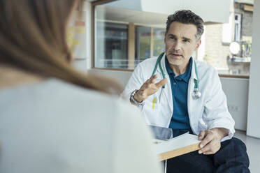 Männlicher Arzt erklärt Patientin im Sitzen im Büro - UUF24475