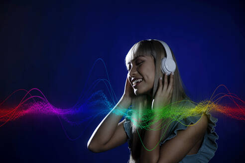 Mehrfarbiges Wellenmuster auf lächelnder junger Frau, die Musik auf blauem Hintergrund hört - EIF01912