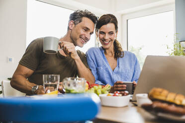 Smiling couple looking at laptop while having breakfast at home - UUF24486