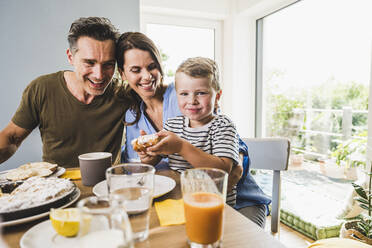 Vater und Mutter betrachten ihren Sohn, der einen Kuchen hält, während sie zu Hause frühstücken - UUF24513