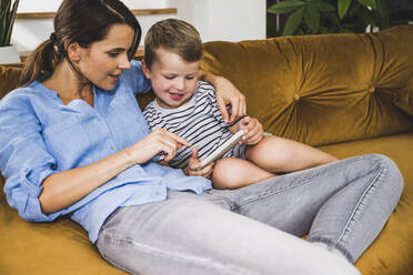 Frau sitzt mit ihrem Sohn auf dem heimischen Sofa und benutzt ein Smartphone - UUF24524