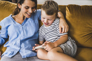 Frau mit Arm um Sohn, der auf dem Sofa zu Hause sein Smartphone benutzt - UUF24525