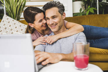 Lächelnde Frau, die einen Mann umarmt, der ein digitales Tablet zu Hause benutzt - UUF24539