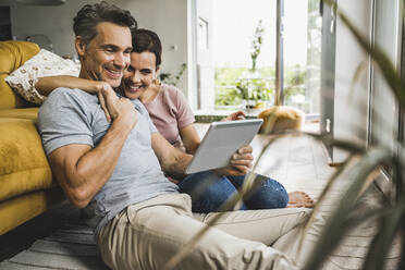 Lächelnde Frau mit einem Arm um einen Mann, der auf ein digitales Tablet schaut, während er zu Hause sitzt - UUF24554