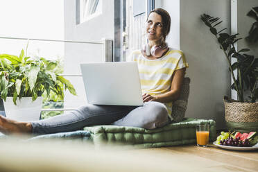 Nachdenkliche Frau, die einen Laptop benutzt, während sie zu Hause sitzt - UUF24559