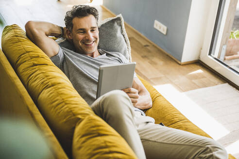 Lächelnder Mann, der ein digitales Tablet benutzt, während er zu Hause auf dem Sofa liegt - UUF24572