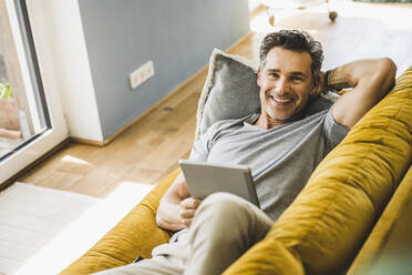 Man relaxing on sofa holding digital tablet at home - UUF24573
