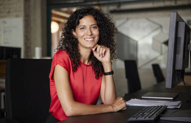 Lächelnde weibliche Fachkraft mit Hand am Kinn, die am Schreibtisch sitzt - RBF08216