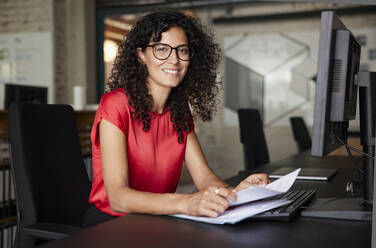 Female professional wearing eyeglasses sitting with documents at desk in office - RBF08220