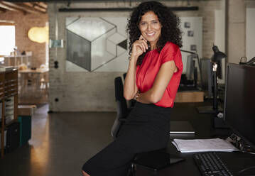 Lächelnde weibliche Fachkraft mit Hand am Kinn am Schreibtisch - RBF08225