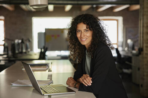 Lächelnde Geschäftsfrau mit Laptop, die sich im Büro auf den Schreibtisch stützt - RBF08239