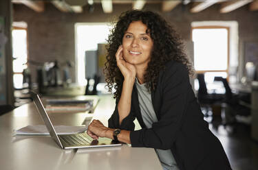 Businesswoman with hand on chin leaning by laptop in office - RBF08240