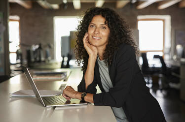 Geschäftsfrau mit Hand am Kinn, die sich im Büro an einen Laptop lehnt - RBF08240