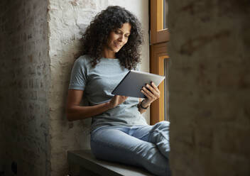 Geschäftsfrau, die ein digitales Tablet benutzt, während sie auf der Fensterbank am Arbeitsplatz sitzt - RBF08246