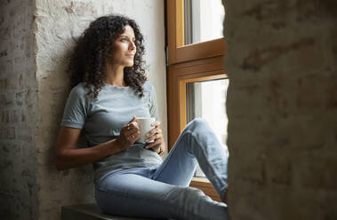 Nachdenkliche Frau hält eine Kaffeetasse, während sie am Fenster sitzt - RBF08247