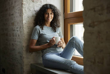 Smiling female professional holding coffee cup while sitting on window sill - RBF08248