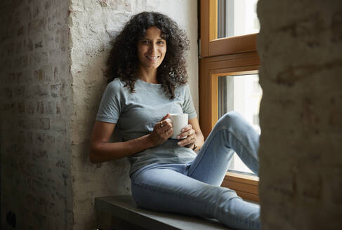 Lächelnde weibliche Fachkraft mit Kaffeetasse auf der Fensterbank sitzend - RBF08248