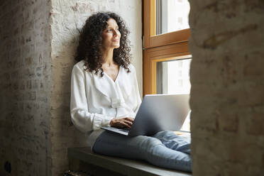 Businesswoman day dreaming while sitting with laptop on window sill - RBF08249